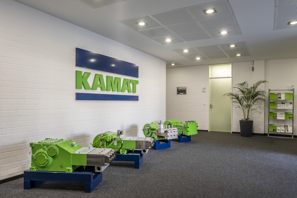 KAMAT plant I Entrance, a hall with the company'S logo at the wall, in front of the wall four green plunger pumps older generations
